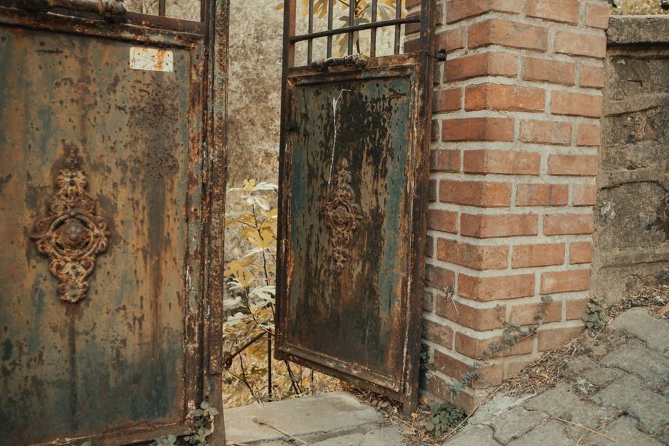 Rusted iron gate doors with ornamental medallions standing open beside a red brick wall.