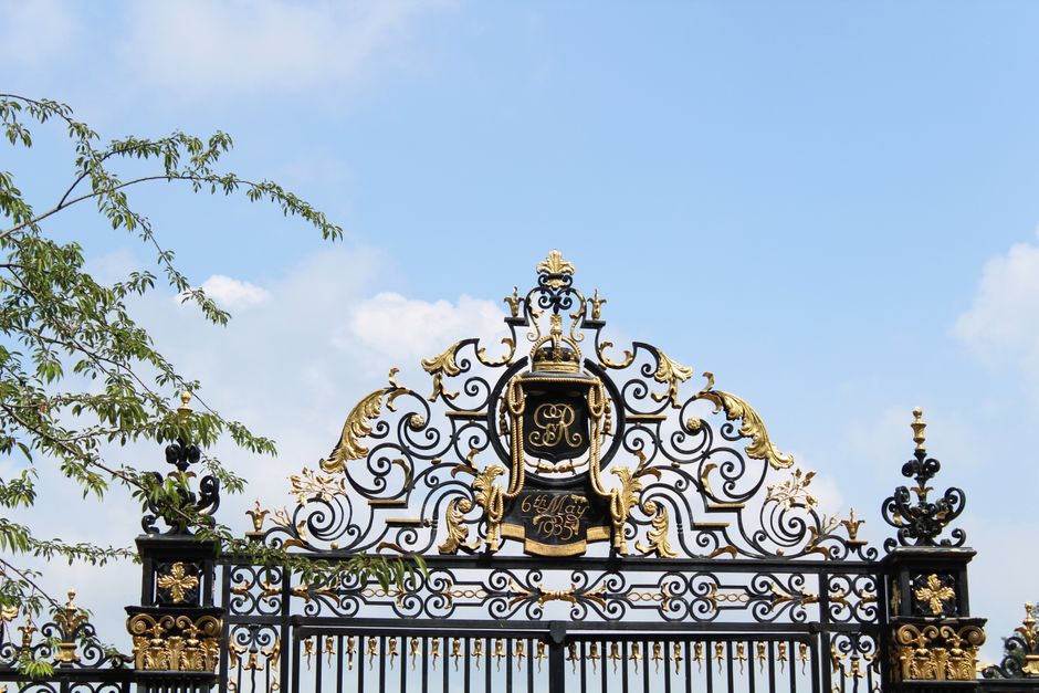 Black and gold wrought iron gate crest with scrolls and finials against a blue sky.