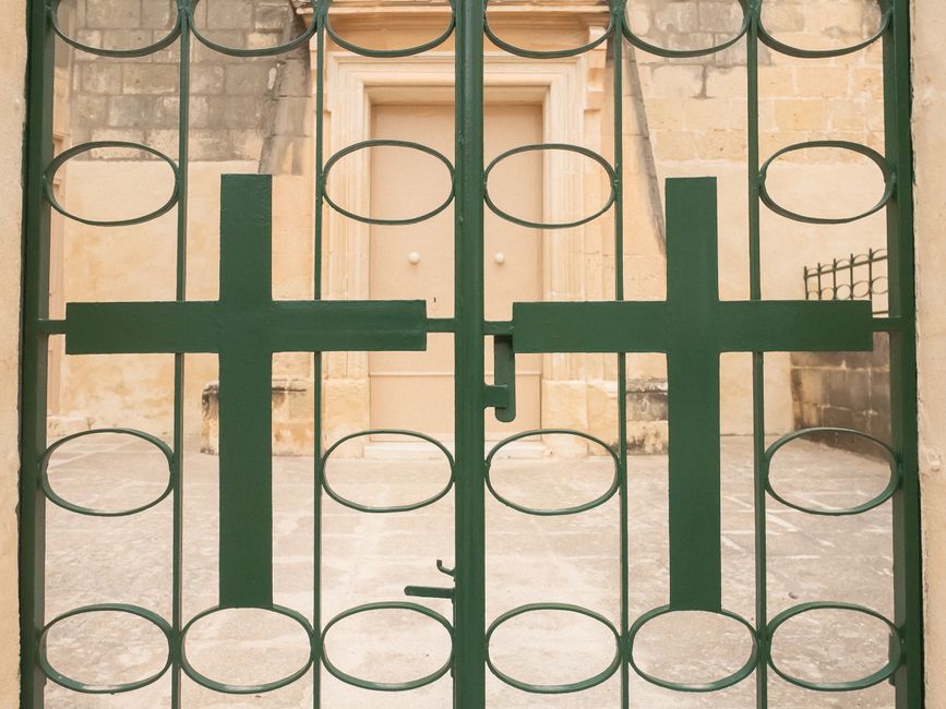 Green painted iron gate with oval cutouts and two cross shapes in front of a stone building.