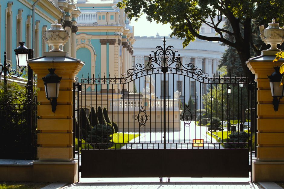 Black wrought iron gate with a scrolled top between two stone pillars with wall lamps.