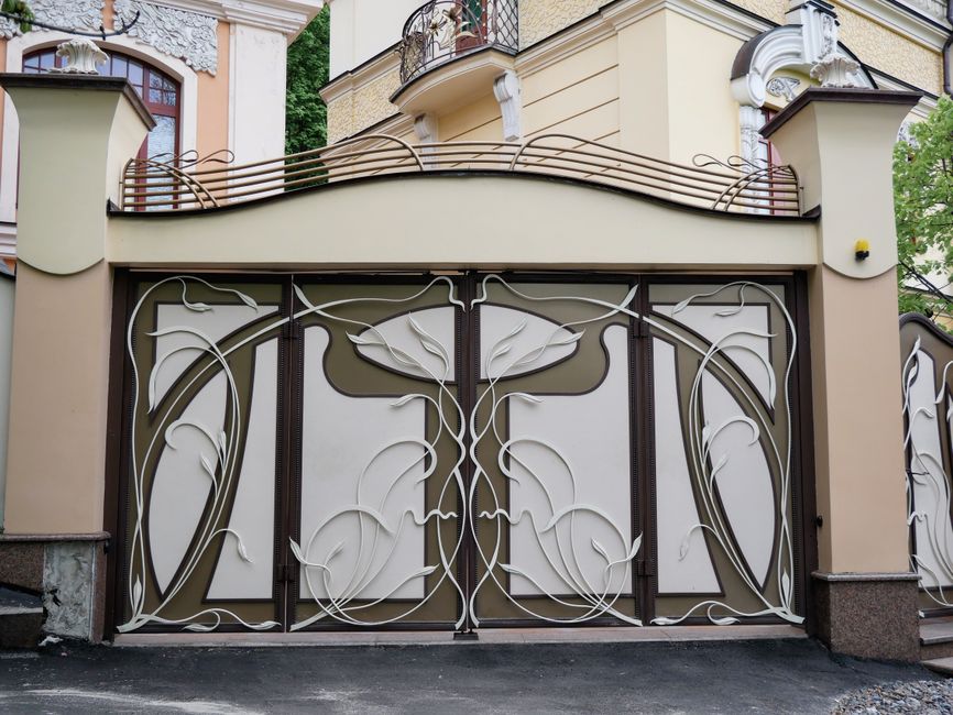 Double leaf iron driveway gate with white floral scrollwork between stucco pillars.