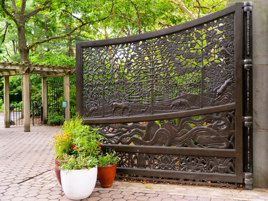 Large curved bronze colored metal gate cast with animal and tree scenes beside potted plants.