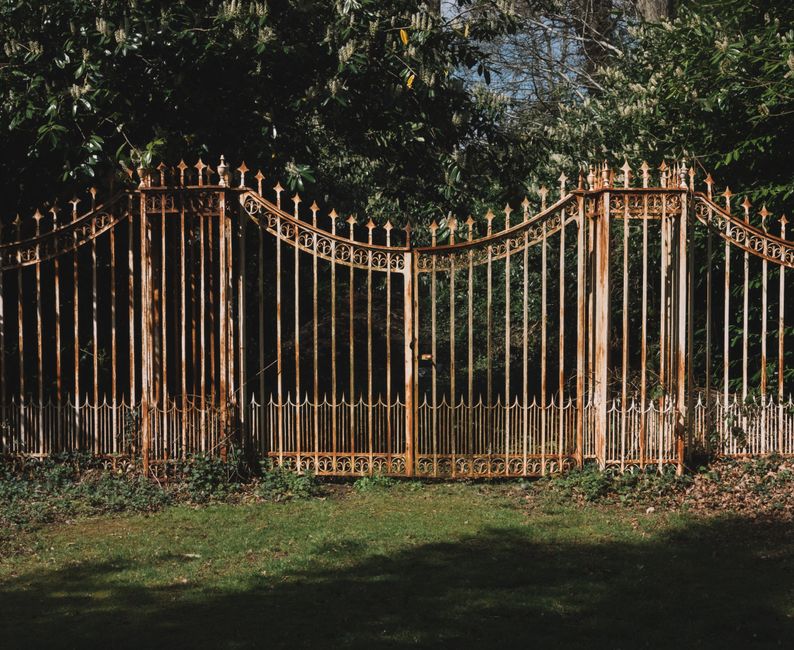 Weathered white painted iron gate with spear-tipped pickets closed across a wooded lawn.